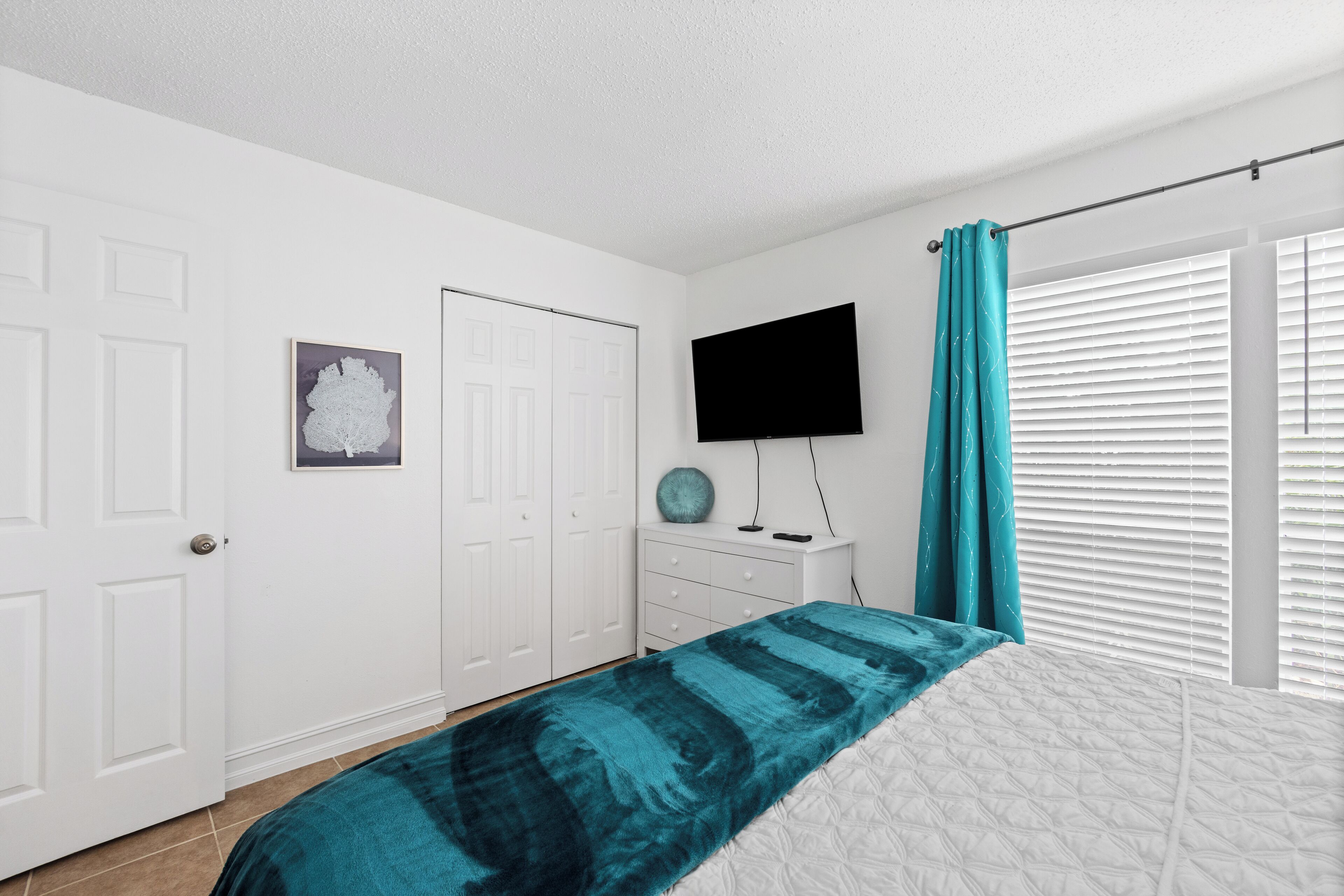 A bedroom with a bed, a TV, a door, and a window with blinds.