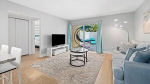 Ein modernes Wohnzimmer mit Fernseher, einer Essecke mit Tisch und Stühlen, einer Couch und einem großen Fenster mit Vorhängen.