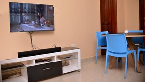 Un área de comedor con un televisor montado en la pared, un mueble blanco de entretenimiento con televisor de pantalla plana y sillas azules alrededor de una mesa de madera.