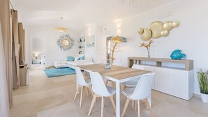 A modern living room with a wooden dining table, white chairs, and a sofa. There is a decorative wall piece and a potted plant on the right side. - Villa with beautiful sea view (Tertenia)