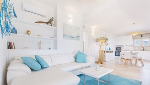A modern living room with a white sofa, blue cushions, a coffee table, and a blue rug. There is a kitchen area with white cabinets and a dining table. - Villa with beautiful sea view (Tertenia)