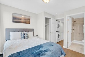 A bedroom with a bed, a nightstand, a washing machine, and a shower area.