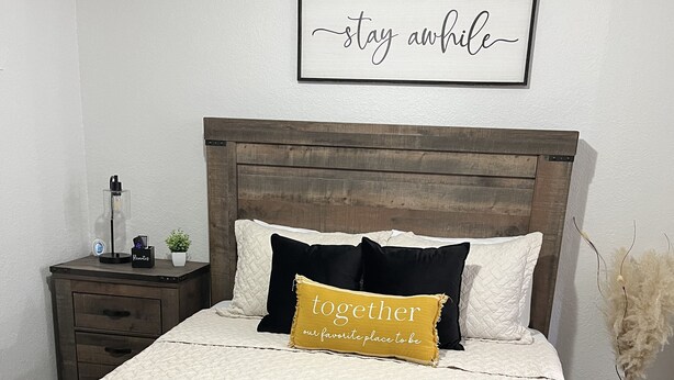 A neatly made bed with a quilted white and black bedspread, a wooden headboard, and a framed sign above reading "stay awhile". - Escape By The Bay (Tampa)