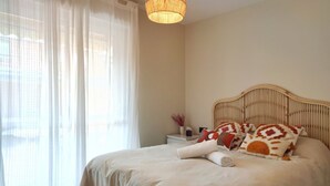 Un lit bien fait, agrémenté d’oreillers à motifs, une table de chevet avec une lampe et des rideaux légers à la fenêtre.