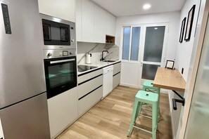 Cocina compacta con microondas, horno y lavabo, un pequeño comedor con taburetes y un ventanal con vidrio esmerilado.