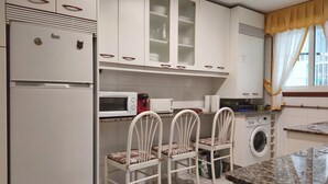 Een keuken met witte kastjes, een magnetron, een wasmachine en een spoelbak. Er staan drie witte stoelen met gekleurde kussens.