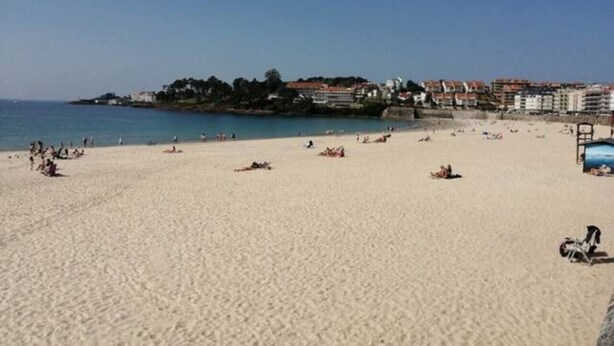 Een strand met mensen die zonnebaden en zwemmen, een heldere lucht en gebouwen op de achtergrond.