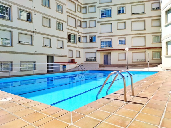 A swimming pool with a tiled deck, surrounded by a multi-story building with balconies and windows.