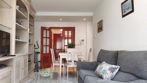 Een woonkamer met een grijze bank, een glazen salontafel en een tv op een kast. Er is een eethoek met een witte tafel en stoelen, en een houten deur die naar een andere kamer leidt.