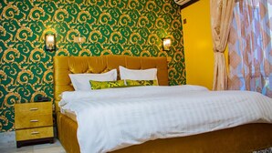 Un letto rifatto con biancheria bianca, un poggiatesta dorato e cuscini decorativi. Un comodino in legno con cassetti e un lampadario fissato al muro.