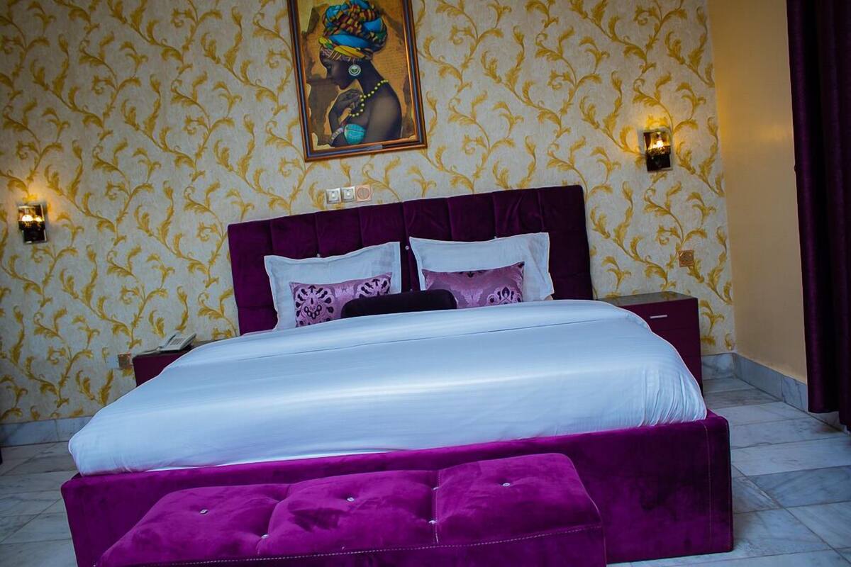 A bedroom with a large bed, purple upholstered headboard, and a matching ottoman. There are two bedside tables with lamps. The walls have a patterned wallpaper.