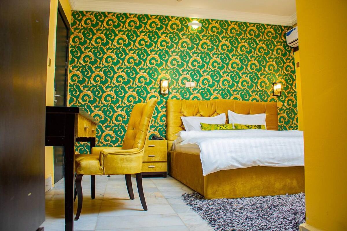 A bedroom with a yellow bed, a yellow chair, and a patterned green and gold wallpaper.