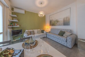 Un salon comprenant un canapé, un téléviseur, une table à manger avec des verres et un saladier rempli de nourriture, ainsi qu’un luminaire suspendu.