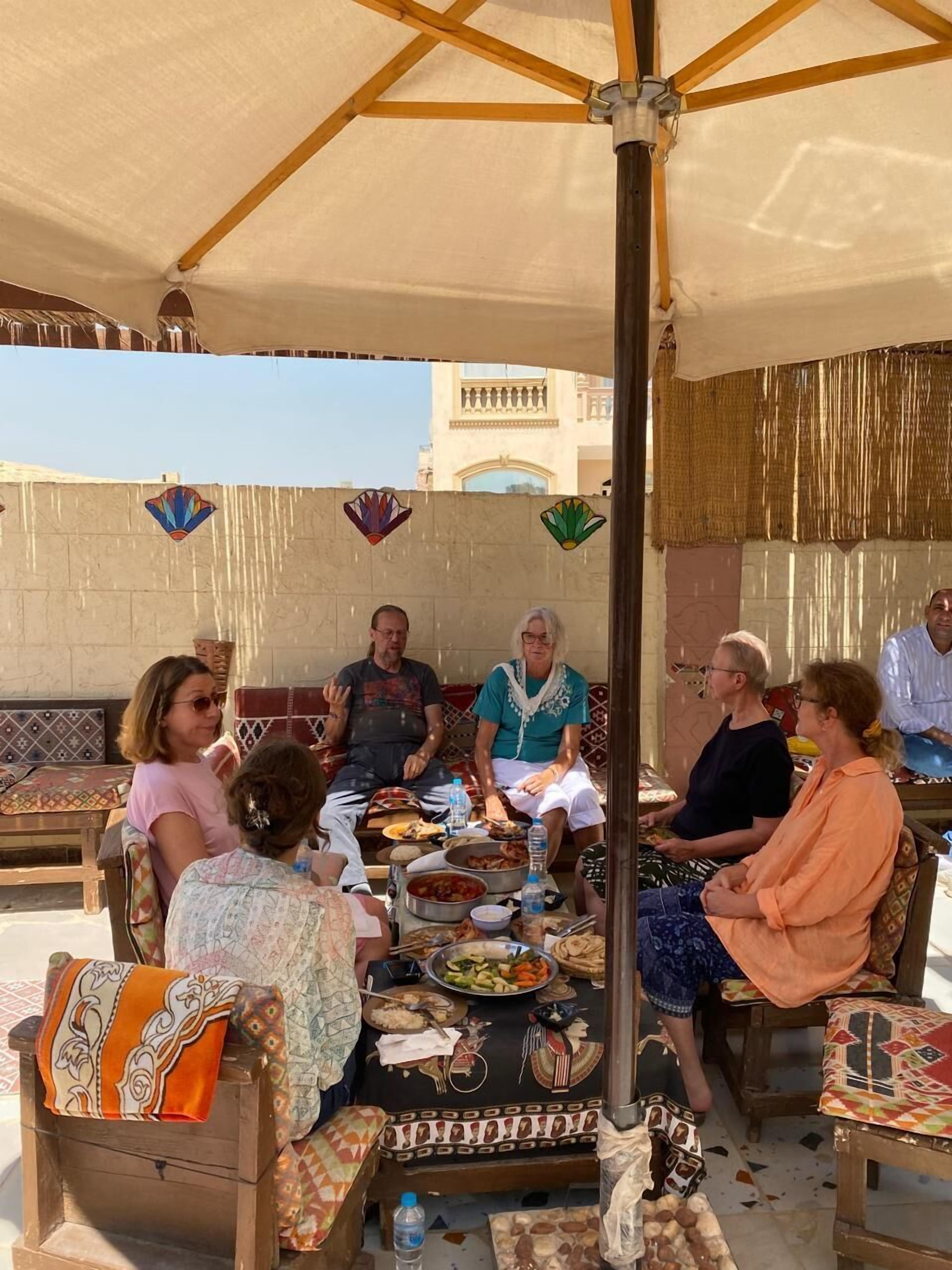 A group of people seated around a table with various dishes, under a canopy with a wooden frame and hanging decorations.