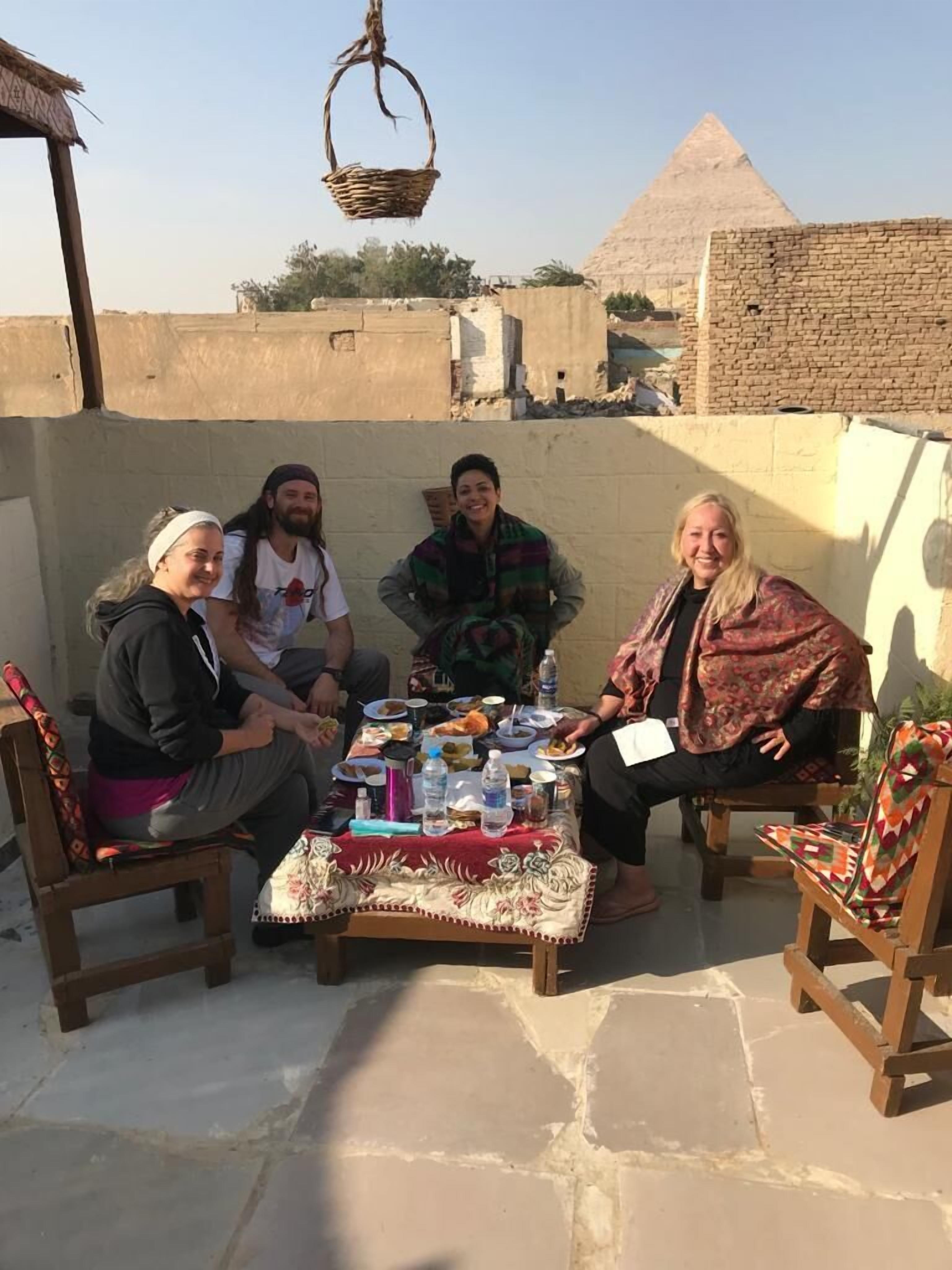 Un gruppo di persone sedute attorno a un tavolo con cibo e bevande, all'aperto con una piramide sullo sfondo.