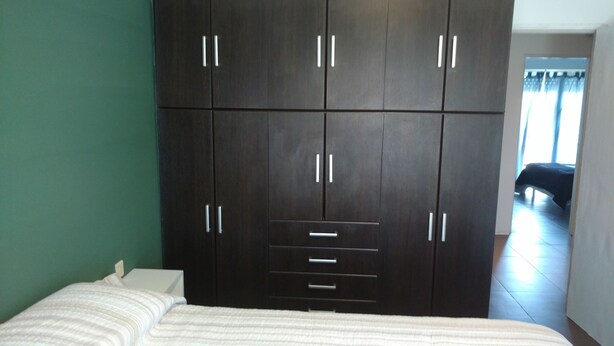 A bedroom with a large dark wooden wardrobe, a bed with a striped white duvet, and a view of a room with a bed through a doorway. - Casa Colinas 2 (Mar del Plata)