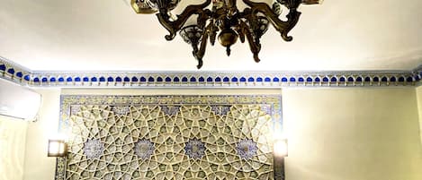 A hotel room with a large bed, two patterned pillows, a wooden headboard, a wall tapestry, and a chandelier.