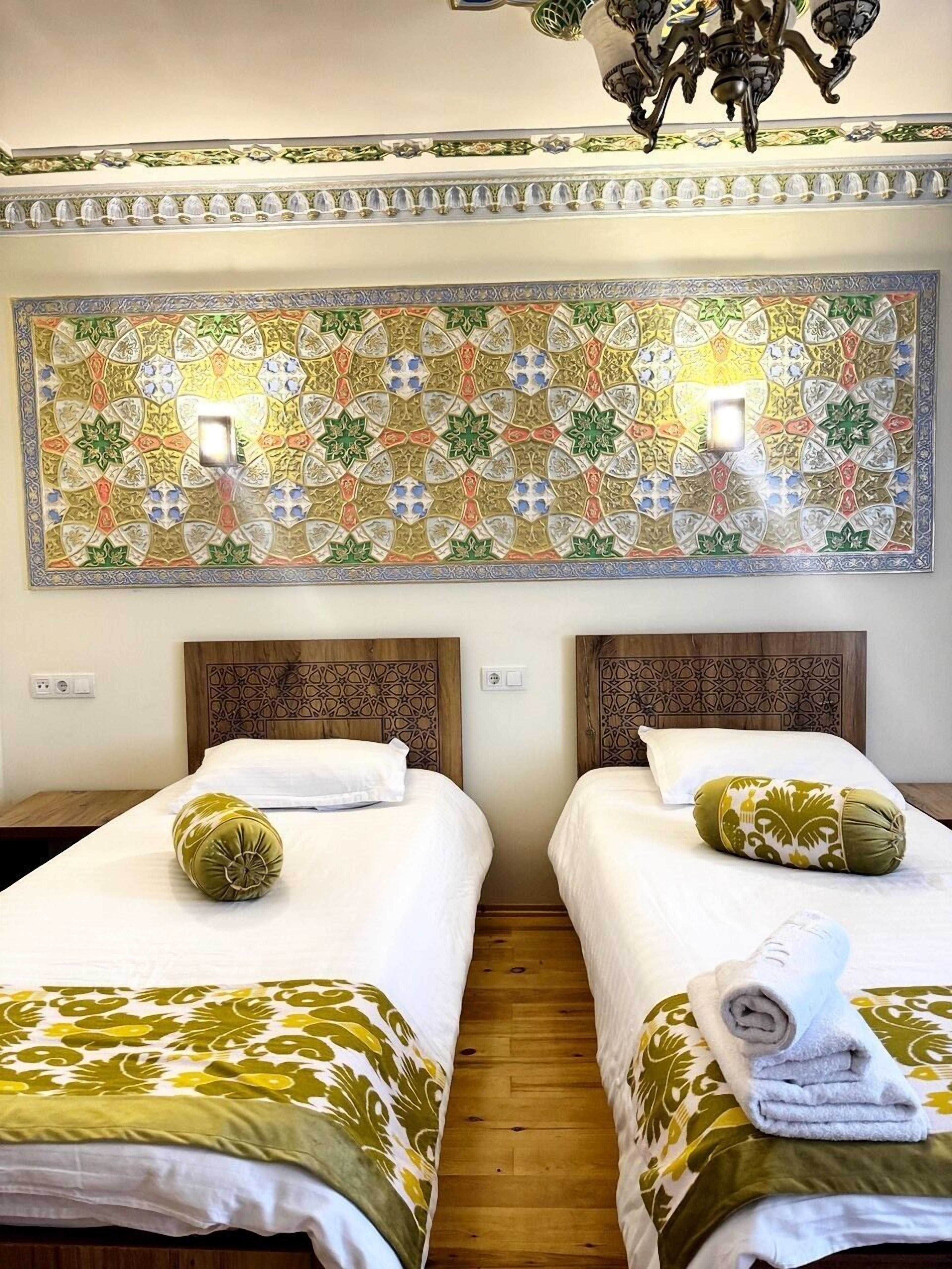 Camera d'albergo con due letti, ognuno con una testiera decorata e un cuscino verde. Sopra i letti si trova un grande pannello decorativo e dal soffitto pende un lampadario.