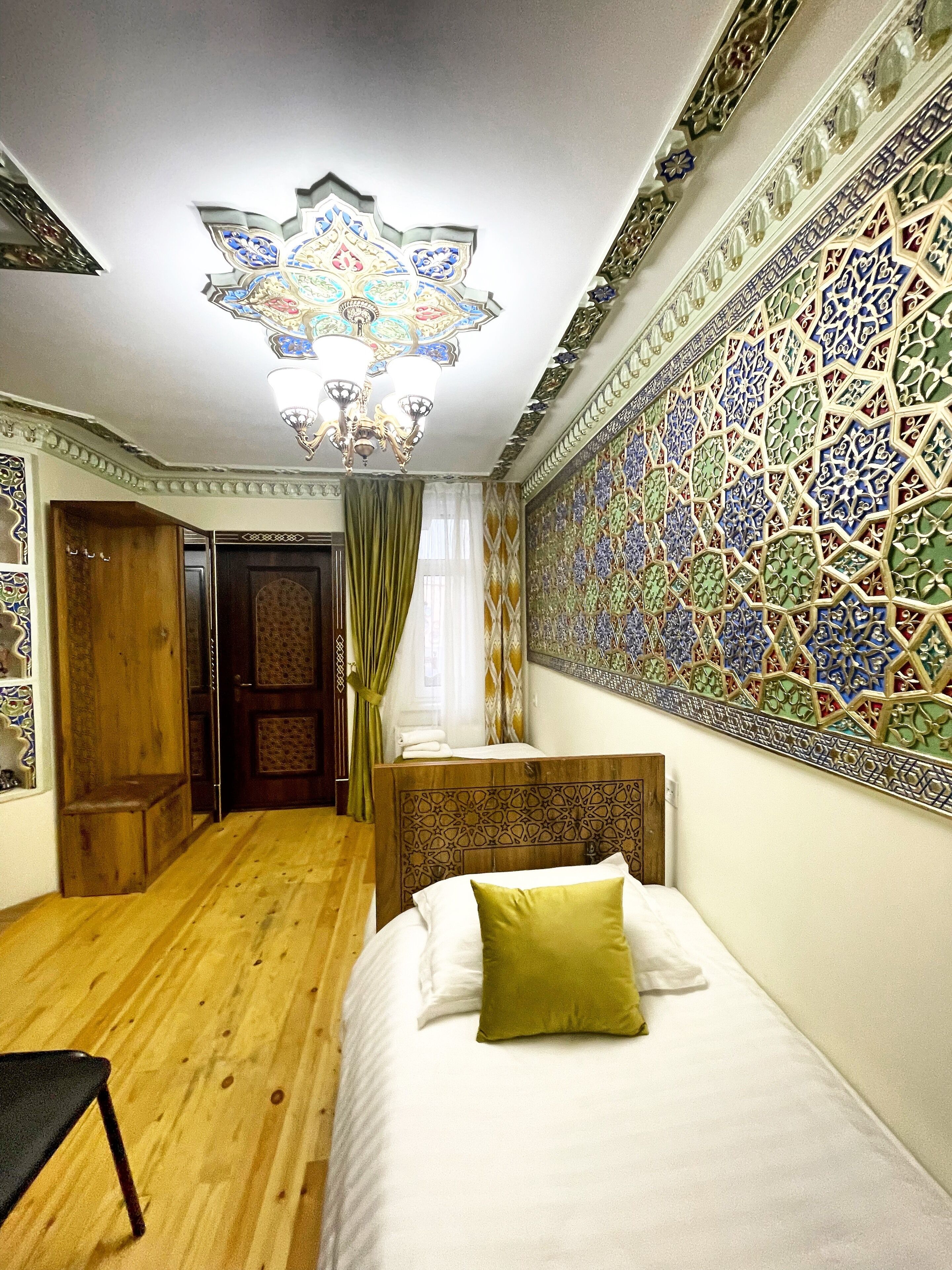 A bedroom with a wooden floor, a bed with a green pillow, a chandelier, and a wall with intricate tile patterns.