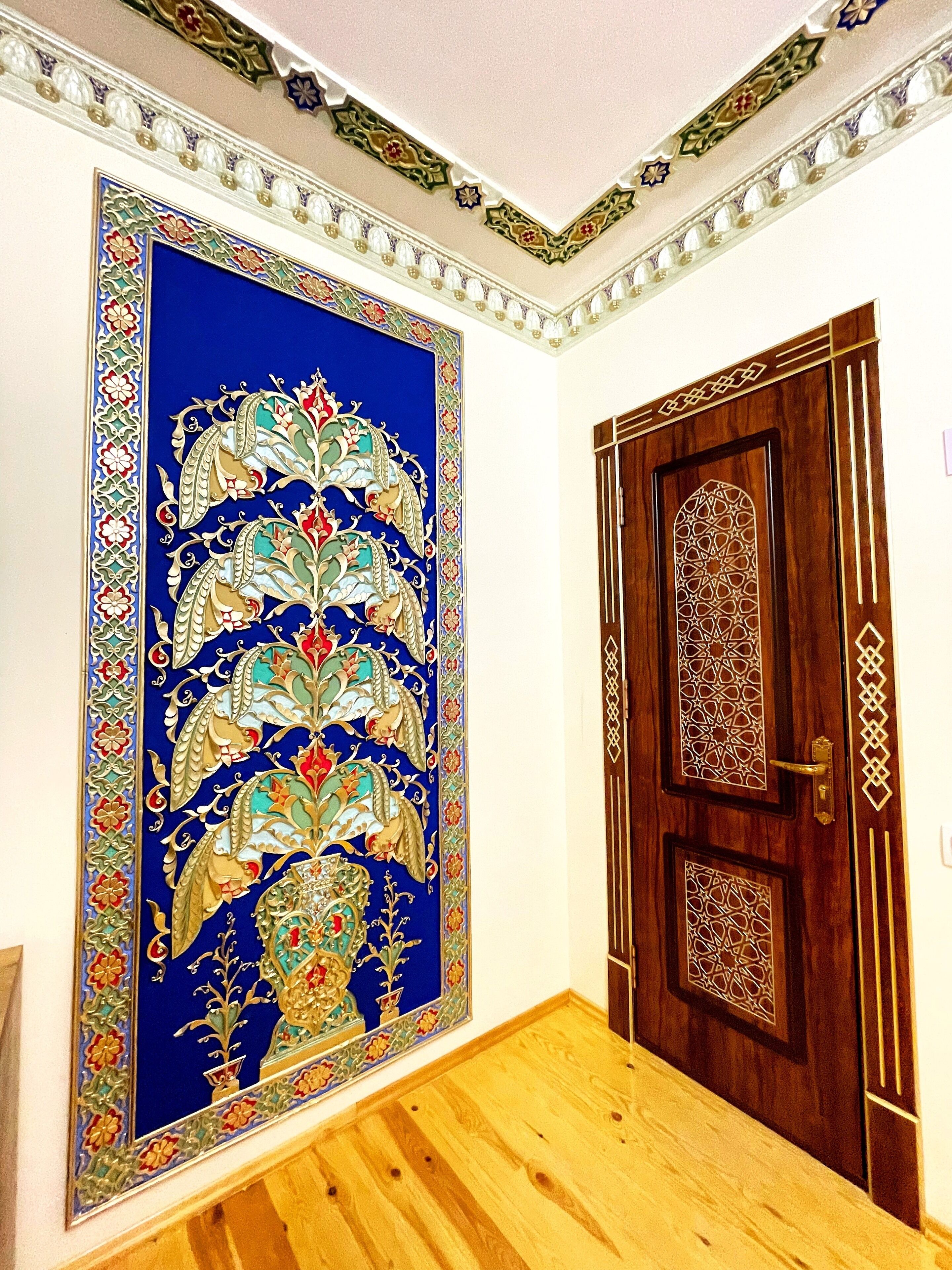 Una habitación con un gran tapiz azul y dorado decorativo en la pared, una puerta de madera con diseños intrincados y suelo de madera.