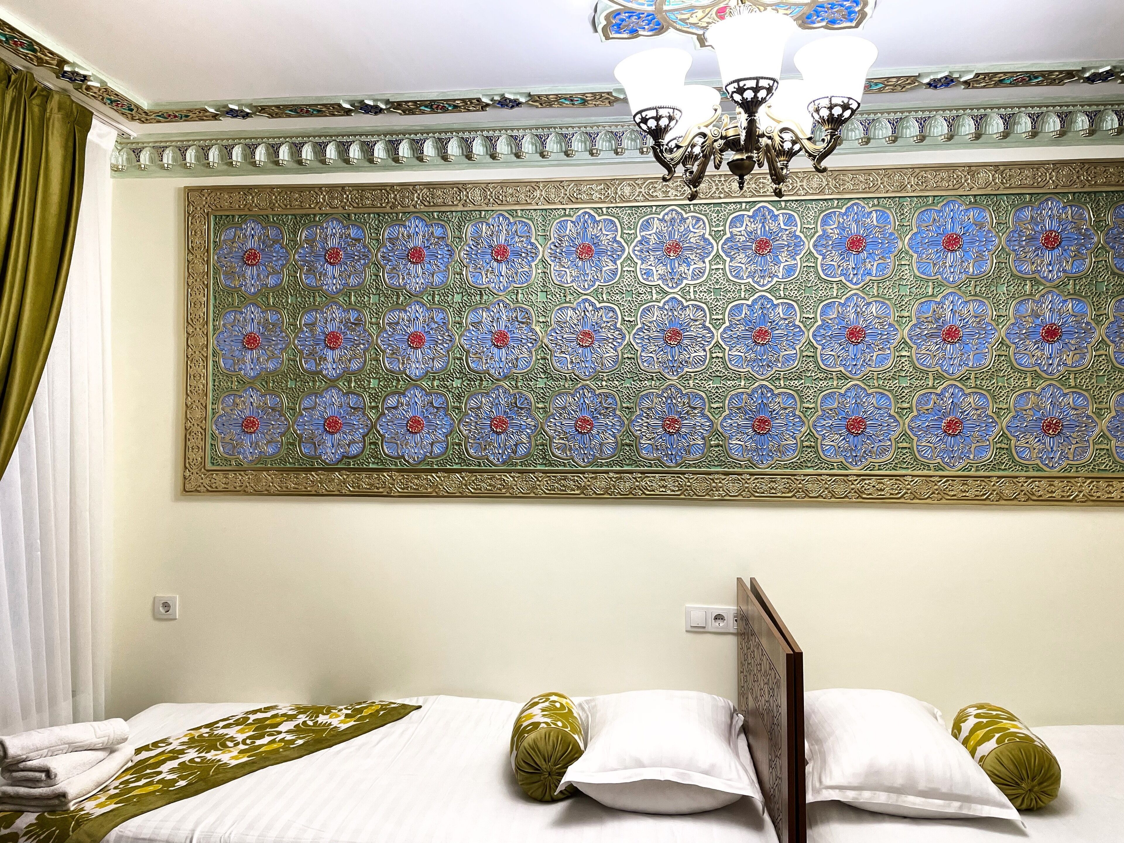 A hotel room with a bed, a decorative wall panel, a chandelier, and a green curtain.
