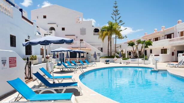 A pool area with blue lounge chairs and umbrellas, surrounded by white buildings with balconies and palm trees. - PORT ROYALE Apartments (Arona)