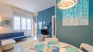 A dining area with a round table set for four, a blue sofa, and a television in the background. - GuestHost - Cozy apartment is located in a strategic position to visit the renowned Ligurian Riviera, suitable for families or groups of friends. Convenient for visiting the nearby towns of the Gulf of Tigullio, such as Chiavari, Santa Margherita and Por (Lavagna)