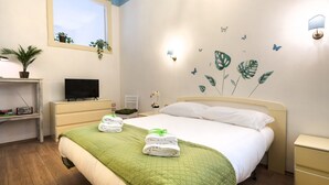 Una cama bien hecha con colcha verde, un televisor sobre un mueble, un escritorio con radio y una pared decorada con motivos de plantas y mariposas.