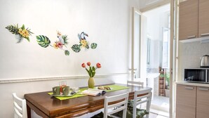 Un comedor con una mesa de madera, sillas blancas, un florero con flores y una pared decorada con arte tropical.