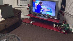 En stue med et fjernsyn på et rødt stativ, en brun sofa og et sofabord i glas.