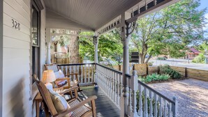 A porch with wooden chairs and a table, a white railing, and a view of a street and trees.