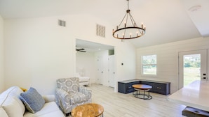 A modern living room with a sofa, patterned armchair, and round coffee table. A kitchen area with dark cabinetry and a dining table is visible.