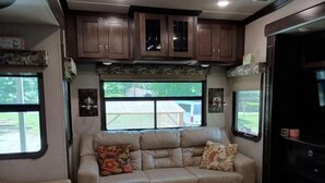 A cozy RV interior with a beige sofa, a window with a view of a house, and wooden cabinets above the sofa. - Arthur's Dream (Hemphill)