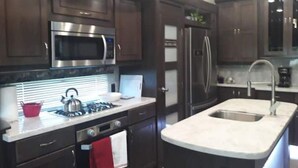 A modern kitchen with dark wood cabinets, stainless steel appliances, a built-in oven, and a large island with a sink. - Arthur's Dream (Hemphill)