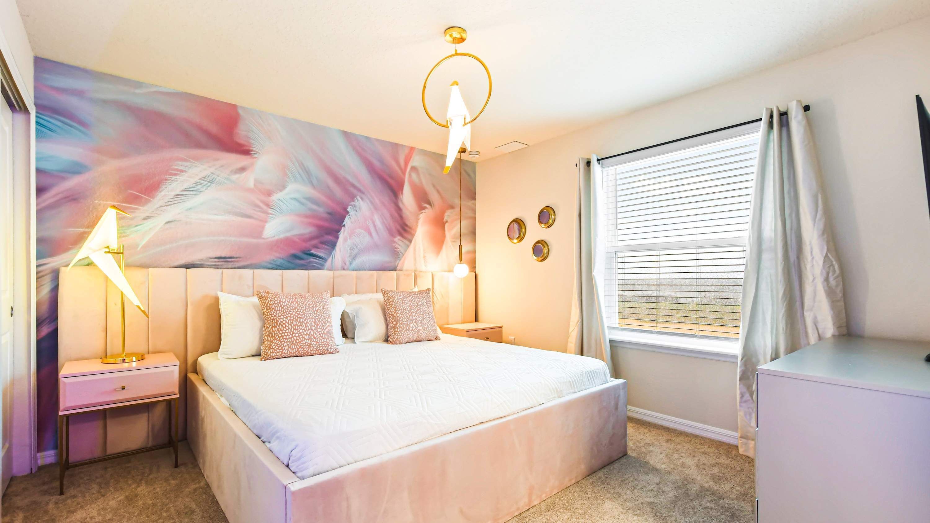 A bedroom with a large bed, bedside tables, a ceiling light fixture, a window with blinds, and a wall with a large abstract painting.