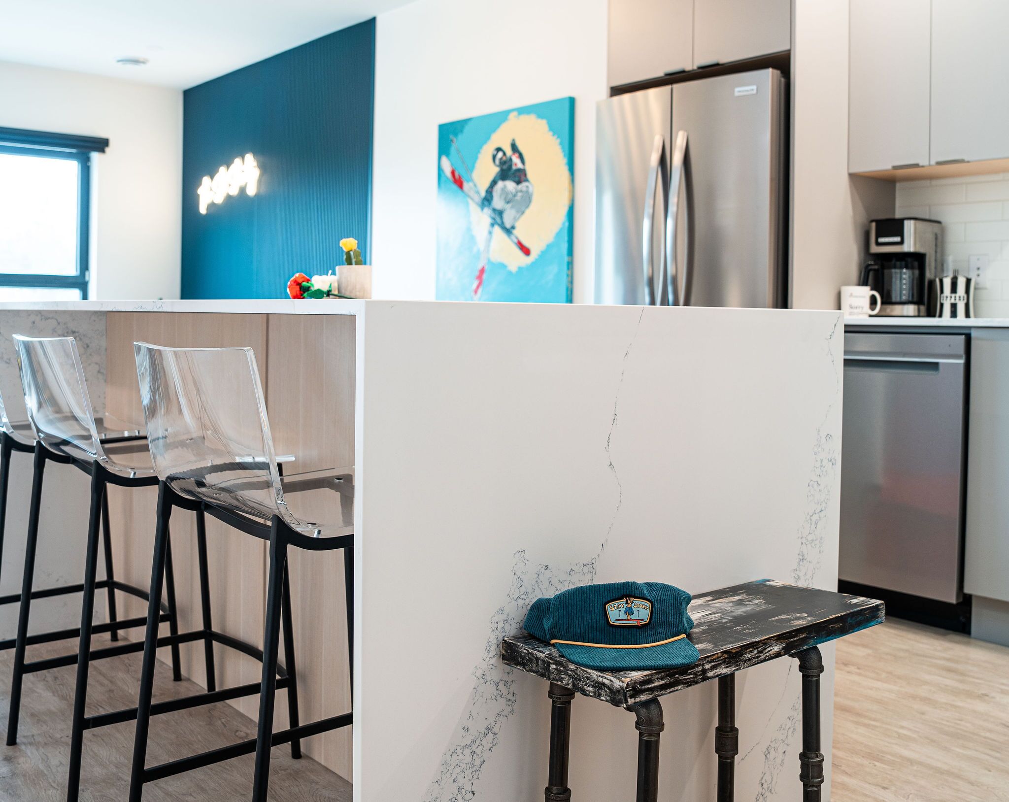 A modern kitchen with a stainless steel refrigerator, a coffee machine, and a dishwasher. There are clear glass chairs and a small table with a hat on it.