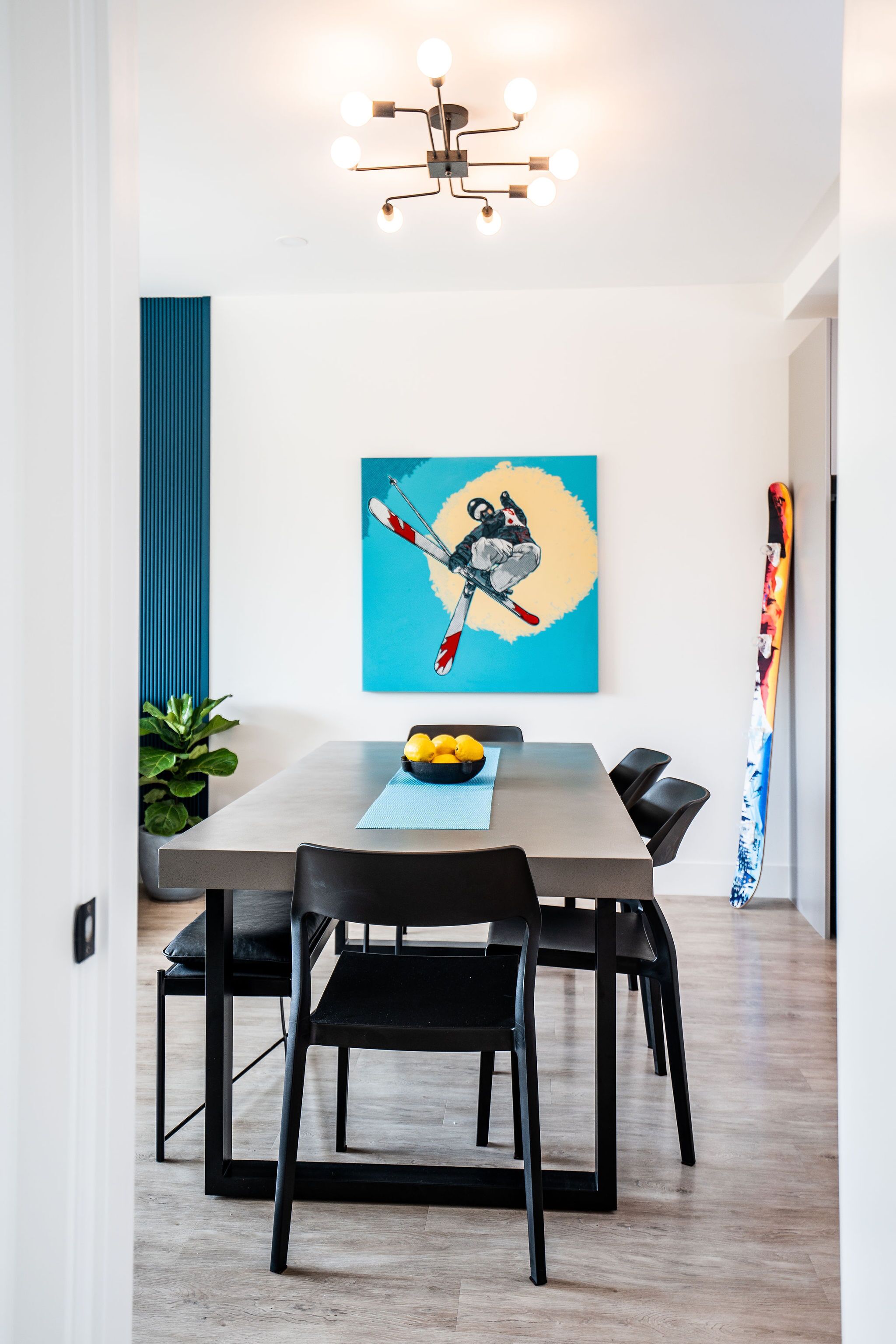 A modern dining area with a wooden table, black chairs, a blue abstract painting, and a ski on the wall.