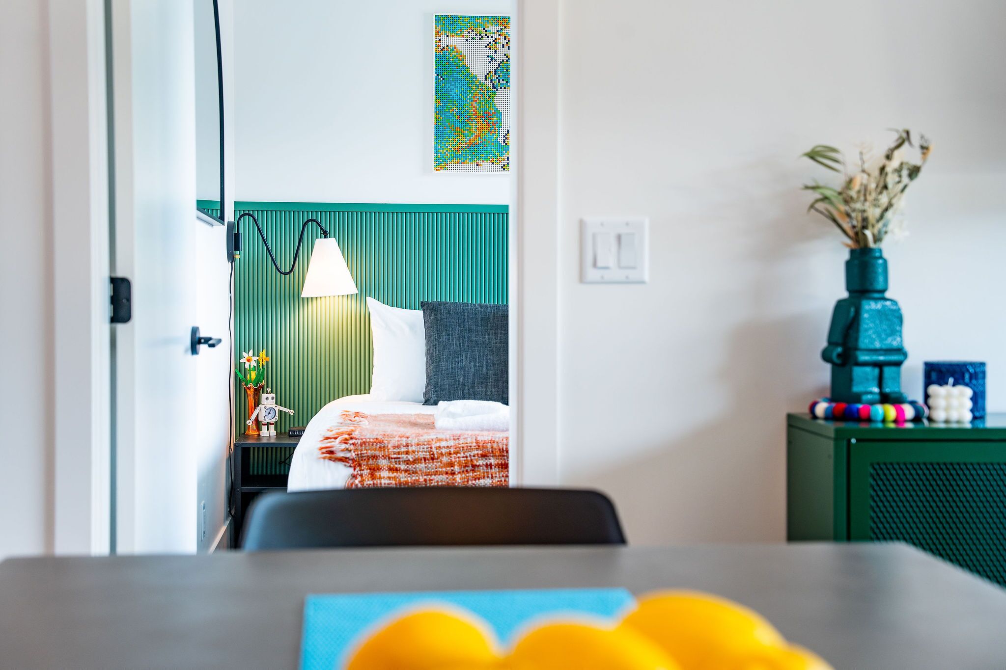 Una habitación de hotel moderna con una cama, una mesita de noche, una lámpara, una obra de arte en la pared y un florero con flores secas.