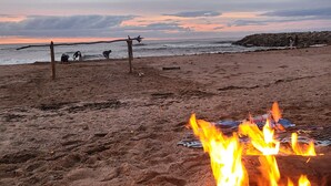 Escena de playa con fogata, costa rocosa y un atardecer.