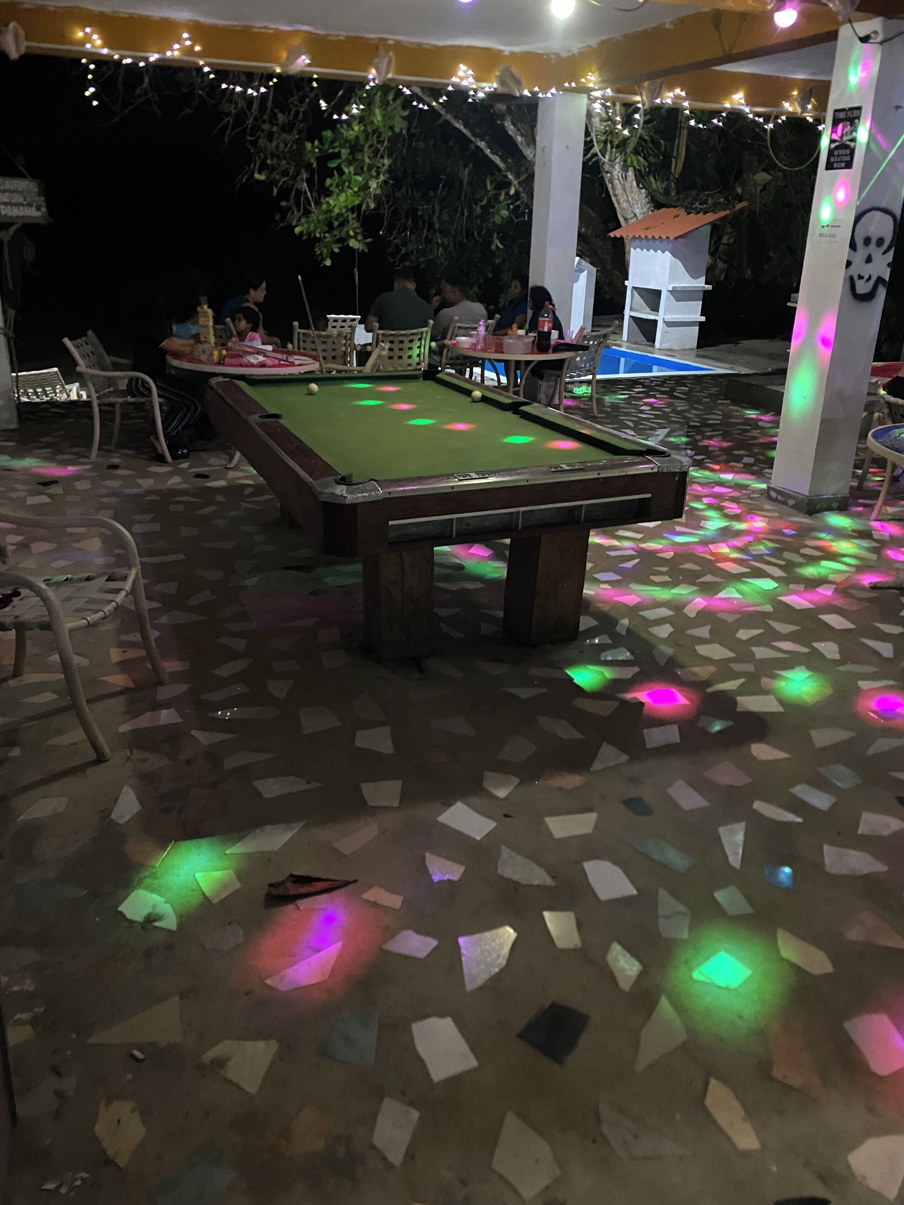 Une table de billard posée sur un sol carrelé, entourée de chaises et de tables, sous une tonnelle illuminée de guirlandes lumineuses.