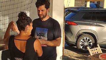 A man and a woman standing in a covered outdoor area with a car parked in the background.