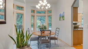 Ein Essbereich mit rundem Tisch, Stühlen und einer Chandelierleuchte. Große Fenster mit vielen Sprossen bieten Blick auf Grünflächen.