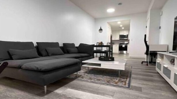 Un salon moderne avec un canapé d’angle gris, une table basse et un tapis. Au fond de la pièce, on aperçoit une cuisine avec des meubles blancs et un coin repas.