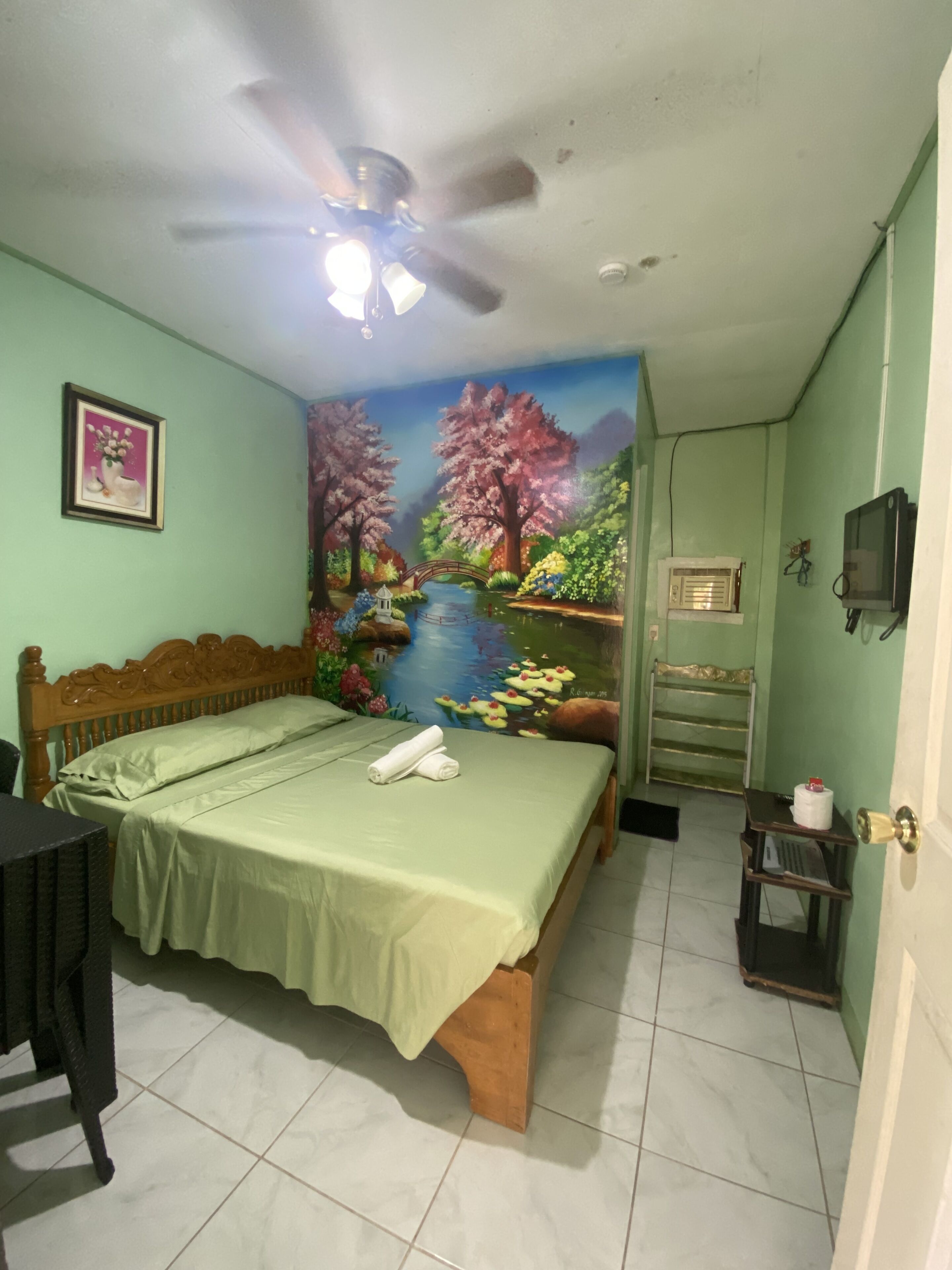Un dormitorio con cama, ventilador de techo, televisión y un cuadro de un paisaje.