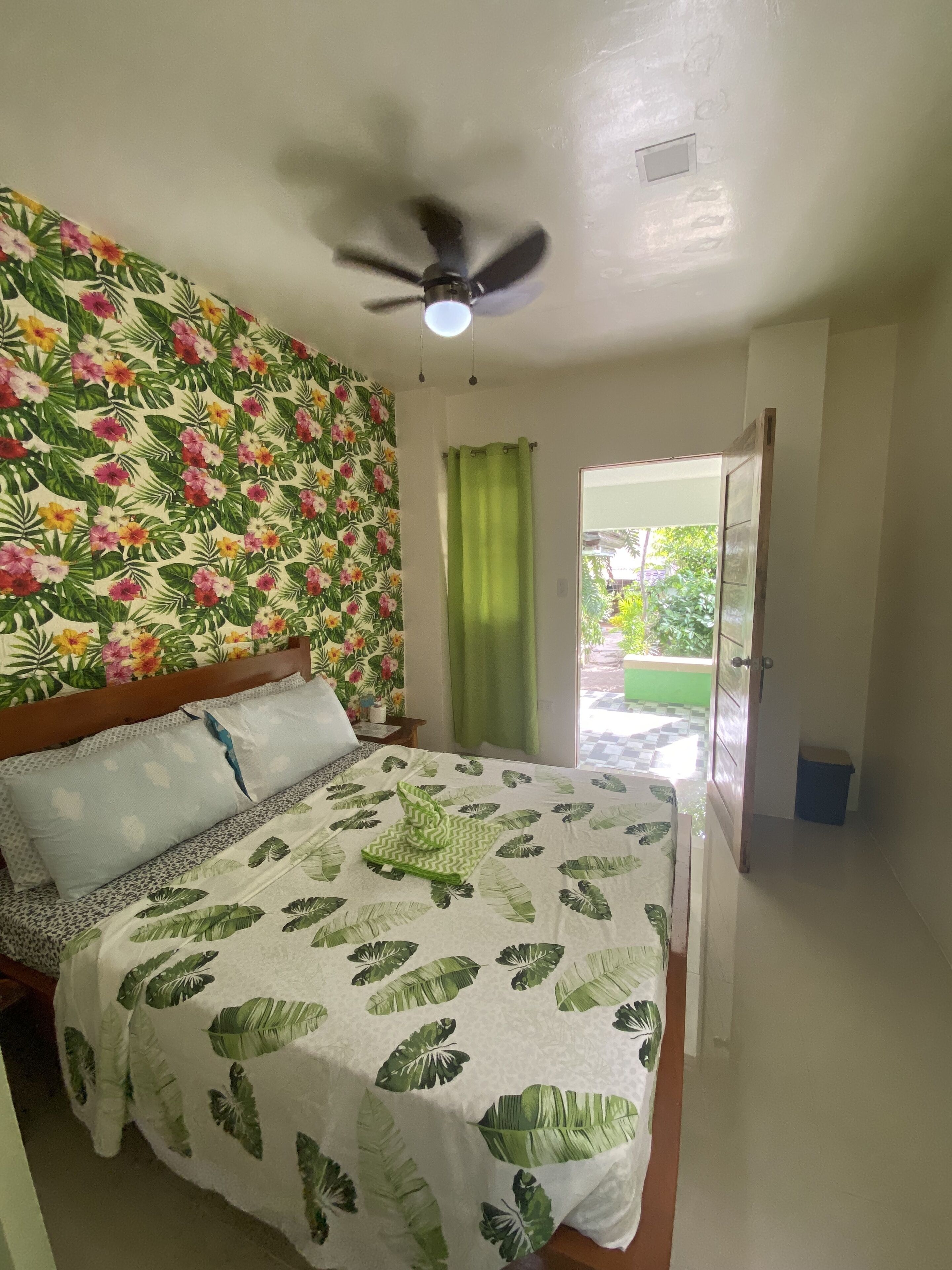 Quarto com cama, ventoinha no teto, cortina verde e vista para o exterior.
