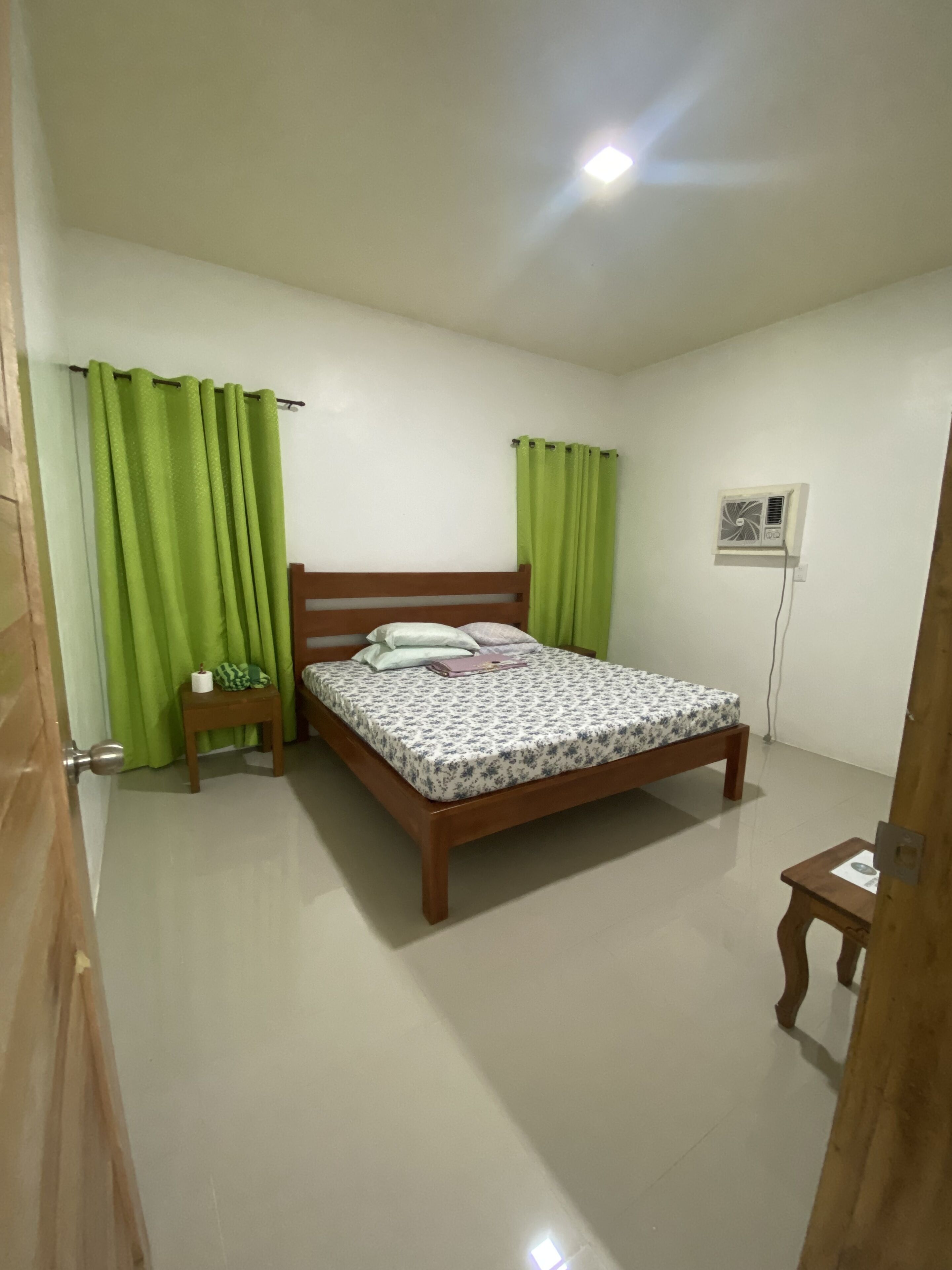 Un dormitorio con una cama de madera, cortinas verdes, una mesita de noche pequeña y un aire acondicionado instalado en la pared.
