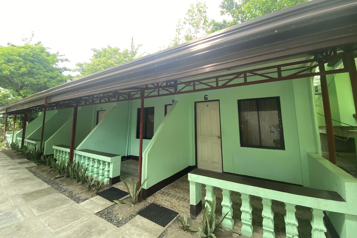 A row of green-roofed buildings with numbered doors, a tiled walkway, and a metal railing.