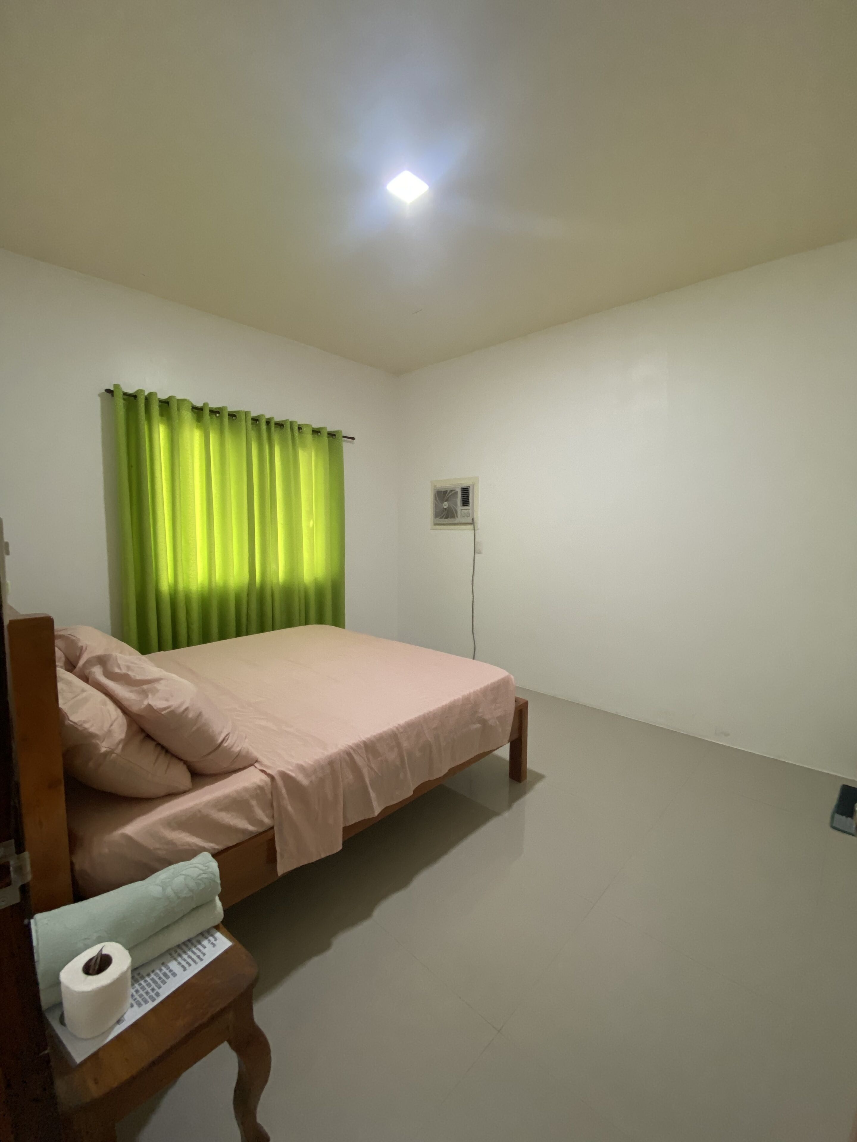 Una habitación sencilla y limpia con una cama, una mesita con un rollo de papel higiénico, una cortina verde y un equipo de aire acondicionado en la pared.