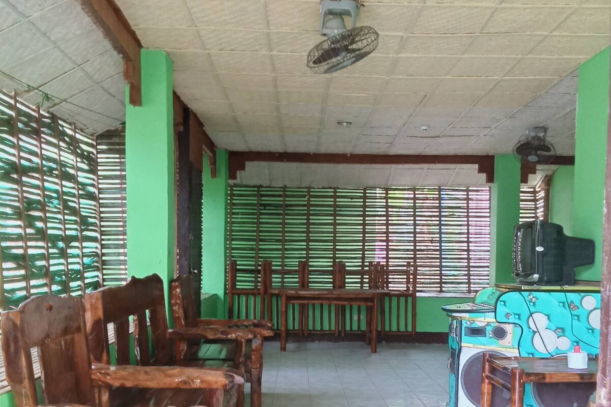A room with wooden chairs and tables, a ceiling fan, and a green partition with windows.