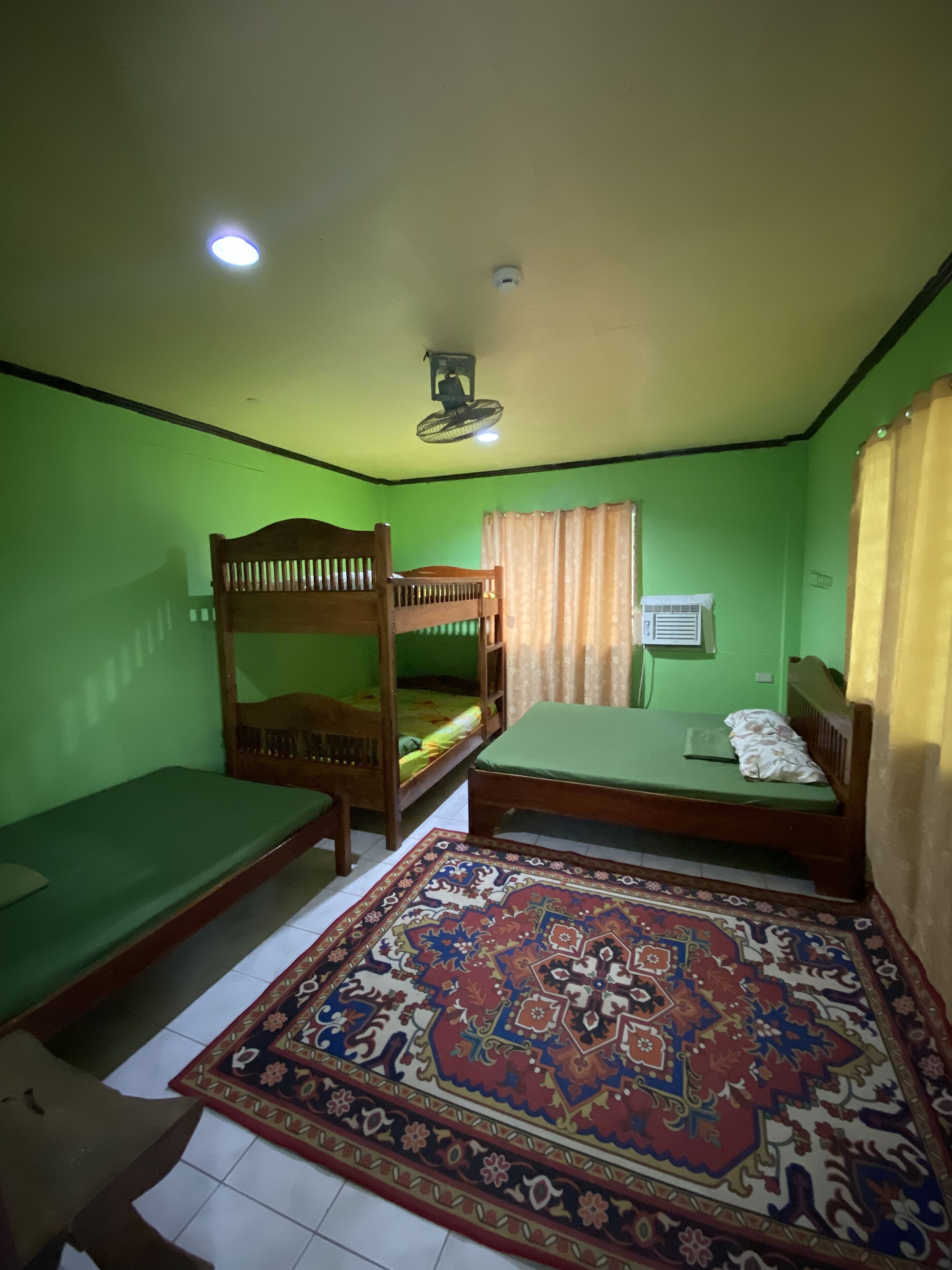 Une chambre avec deux lits superposés, un lit simple, un ventilateur de plafond, une fenêtre avec des rideaux et un tapis à motifs.